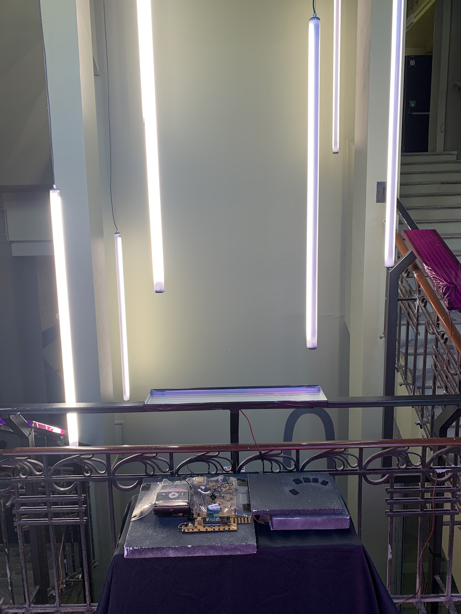 In a stairwell, a control surface and circuit boards on a table. Behind it on the railing is a shiny panel. Hanging behind them in the stairwell are vertical tubular light fixtures.